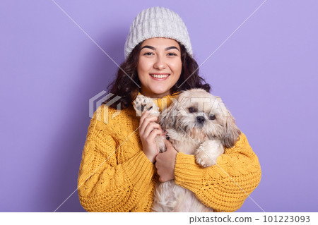 Happy beautiful female with dark wavy hair, carries little fluffy dog, plays and spend time together, posing isolated over lilac studio background, pretty woman wearing yellow knitted sweater and cap. Happy beautiful female with dark wavy hair, carries little fluffy dog, plays and spend time together, posing isolated over lilac studio background, pretty woman wearing yellow knitted sweater and cap. 101223093