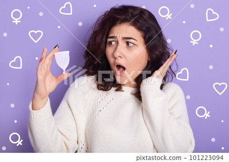 Shocked impressed female with black curly hair having surprised facial expression, holding menstrual cup in one hand, raising one hand, opening mouth widely, posing over lilac background in studio. 101223094