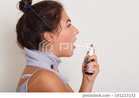 Profile portrait of attractive upset young woman with bun on head, uses spray to treat sore throat, has scarf around neck, catch cold. Throat pain, painful swallowing concept. Copy space for promotion 101223132