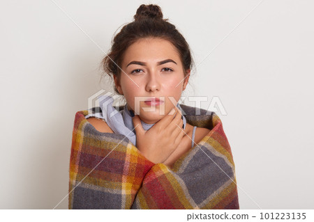 Image of sick young dark haired woman with bun on head, posing isolated over white studio background, unhealthy lady touches neck, wearing checkered plaid, lady has sore throat or catch cold. 101223135