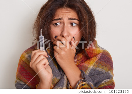 Close up portrit of woman being examined by doctor, needs injection, holding syringe in hands, looks with scared on it, wrapped checkered plaid on shoulders. Healthy care and illness concept. 101223155