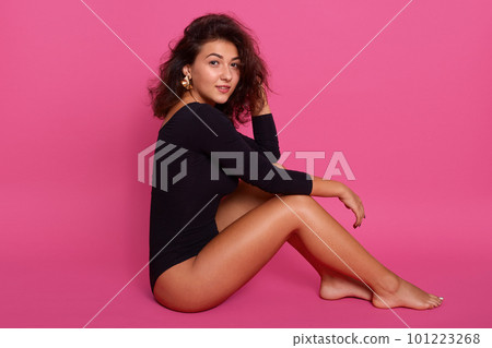 Woman with perfectly shaped body sitting on floor and holding one hand on her leg and touching her dark wavy hair with other, attractive girl dresses black combidress, looks at camera, looks confident 101223268