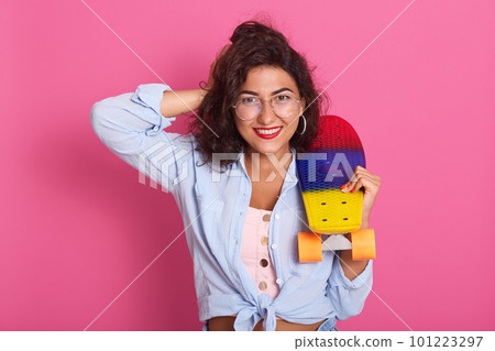 Indoor studio shot of charming energetic lady with bright make up wearing blue shirt, top and accessories, having black curly hair, standing isolated over pink background. People and hobby concept. 101223297