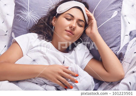 Indoor shot of sleeping girl with blindfold on her head, female wearing white t shirt, lying in bed under blanket, keeps eyes closed, relaxing at home in morning, nowhere to rush during weekend.. 101223501