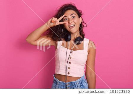 Close up portrait of young woman making victory or peace sign and covering her eye with it, posing over pink wall background indoors, happy girl wearing rosy top and jrans, has heandphones around neck 101223520