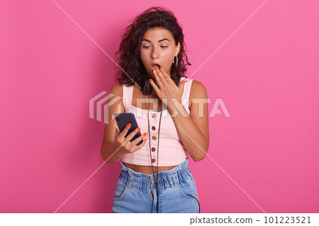 Half length ortrait of young brunette woman in rose t shirt and jeans or short, holding smart phone, looks schocked and surprised, looking at her device with open mouth, isolated on pink background. Half length ortrait of young brunette woman in rose t shirt and jeans or short, holding smart phone, looks schocked and surprised, looking at her device with open mouth, isolated on pink background. 101223521