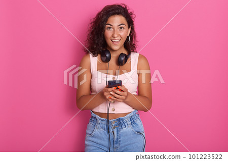 Portrait of happy beautiful young woman holding phone in hand and smiling, wearing casual clothing, posing isolated over pink background, female with dark wavy hair, expressing astonishment. Portrait of happy beautiful young woman holding phone in hand and smiling, wearing casual clothing, posing isolated over pink background, female with dark wavy hair, expressing astonishment. 101223522