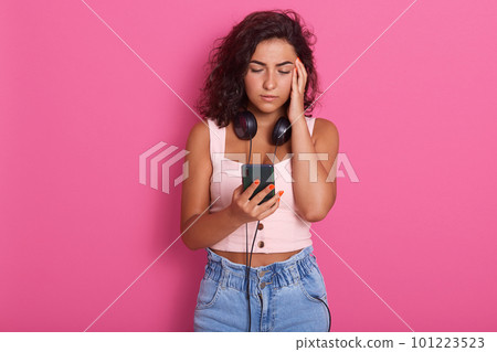 Close up portrait of young sad woman looks at her phone's screen, having headache, adorable female wearing rose top and jeans, having dark hair, keeps finger on temple, isolated over rosy background. Close up portrait of young sad woman looks at her phone's screen, having headache, adorable female wearing rose top and jeans, having dark hair, keeps finger on temple, isolated over rosy background. 101223523