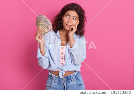 Young attractive smiling woman thinking how to spend her bunch of money, female wearing blue shirt and jeans, standing against pink wall with pensive expression. Young attractive smiling woman thinking how to spend her bunch of money, female wearing blue shirt and jeans, standing against pink wall with pensive expression. 101223860