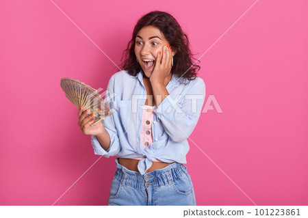 Portrait of shocked young brunette woman wearing casual clothes, female holding fan of cash money, has surprised facial expression, isolated over pink background in studio. People lifestyle concept. 101223861