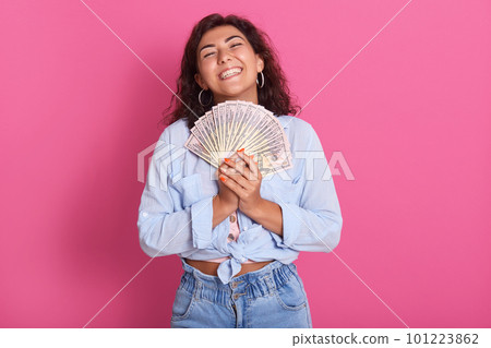 Indoor studio shot of delighted cheerful model posing isolated over bright pink background, closing her eyes with satisfaction, holding many banknotes in both hands. People and money concept. Indoor studio shot of delighted cheerful model posing isolated over bright pink background, closing her eyes with satisfaction, holding many banknotes in both hands. People and money concept. 101223862
