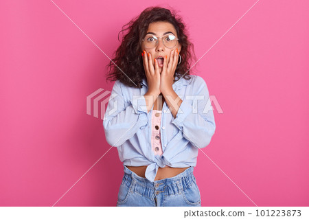 Close up portrait of amazed horrified female keeps mouth opened and hands on cheeks, being shocked, pretty lady expresses great surprisment, looking at camera, isolated over rose background. Close up portrait of amazed horrified female keeps mouth opened and hands on cheeks, being shocked, pretty lady expresses great surprisment, looking at camera, isolated over rose background. 101223873