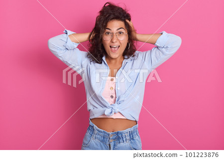 Horiontal shot of shouting, screaming young woman keeps hands on head, female dressed blue shirt and spectacles, studio portrait of adorable exited girl. Emotions, feelings and states of mind concept. 101223876