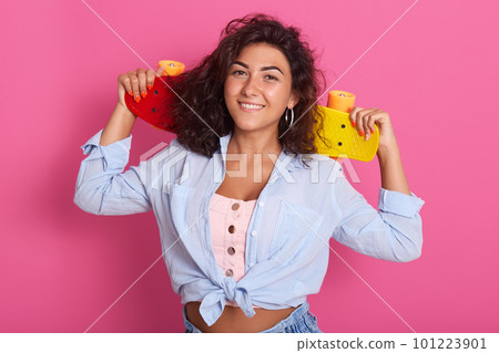 Image of cheerful good looking female with dark curly hair smiling sincerely, having pleasant facial expression, holding her colorful skateboard in hands aback, being in high spirits. Mood concept. Image of cheerful good looking female with dark curly hair smiling sincerely, having pleasant facial expression, holding her colorful skateboard in hands aback, being in high spirits. Mood concept. 101223901
