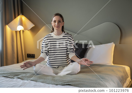 Young woman meditating with closed eyes in bright bedroom, sitting on bed 101223976