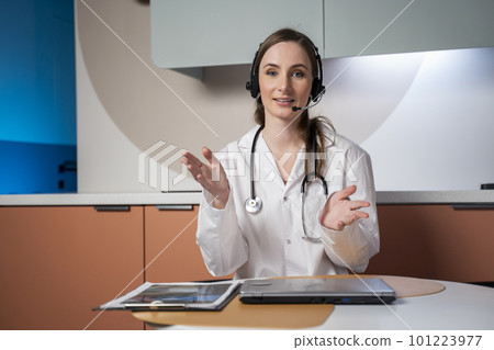 Young female doctor in a white medical uniform with a stethoscope, using a computer laptop, talks via videoconference with a patient, looking into the camera at a healthcare hospital 101223977