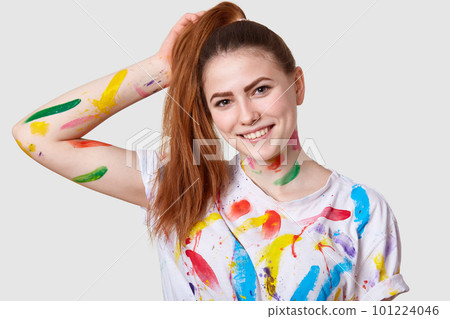 Headshot of pleased cheerful woman has dark hair combed in pony tail, smiles gently, has dirty body and t shirt with acrylic paints, brings creativity to life, isolated over white studio wall Headshot of pleased cheerful woman has dark hair combed in pony tail, smiles gently, has dirty body and t shirt with acrylic paints, brings creativity to life, isolated over white studio wall 101224046