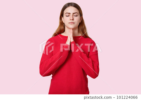 Photo of spiritual woman keeps eyes closed, hands in praying gesture, dressed in red outfit, prays for good wellness, poses over light pink background. People, faith and spirituality concept Photo of spiritual woman keeps eyes closed, hands in praying gesture, dressed in red outfit, prays for good wellness, poses over light pink background. People, faith and spirituality concept 101224066