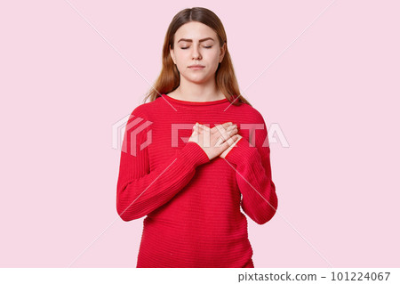 Studio portrait of restful impressed woman keeps both hands near heart, feels touched to hear heart piercing story, expresses good feelings, closes eyes, dressed in red jumper, isolated over rosy wall 101224067