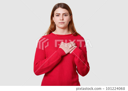 Calm dark haired female with serious facial expression, keeps both palms on heart, being touched by something, dressed in casual red sweater, isolated over white background. Body language concept Calm dark haired female with serious facial expression, keeps both palms on heart, being touched by something, dressed in casual red sweater, isolated over white background. Body language concept 101224068