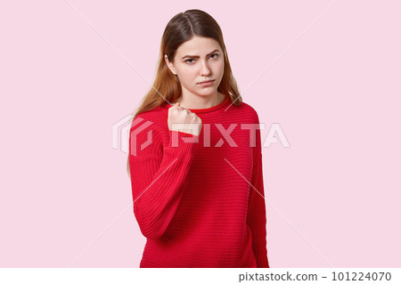 Photo of sad angry young European woman with straight dark hair, shows fist at camera, dressed in red jumper, poses over rosy background, expresses threating, reproaches and promises to revenge. Photo of sad angry young European woman with straight dark hair, shows fist at camera, dressed in red jumper, poses over rosy background, expresses threating, reproaches and promises to revenge. 101224070