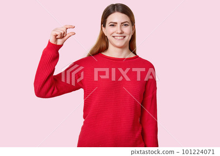 Horizontal shot of attractive young woman shows tiny object, gestures with hands, dressed in red clothes, has toothy smile, demonstrates how much she recieves money, models over rosy background Horizontal shot of attractive young woman shows tiny object, gestures with hands, dressed in red clothes, has toothy smile, demonstrates how much she recieves money, models over rosy background 101224071