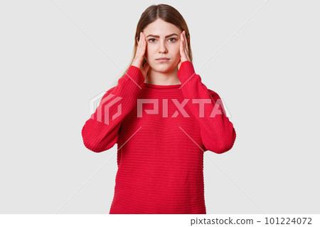 Studio shot of displeased female suffers from headache, dressed in red sweater, keeps hands on temples, has upset facial expression, feels bad, isolated over white background. Healthy care concept. Studio shot of displeased female suffers from headache, dressed in red sweater, keeps hands on temples, has upset facial expression, feels bad, isolated over white background. Healthy care concept. 101224072
