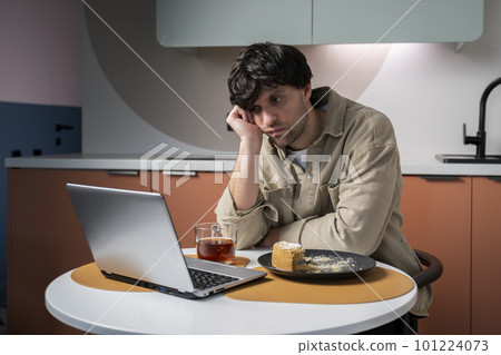 Man eats a cake and drinks tea for breakfast sitting at a table in the kitchen Man eats a cake and drinks tea for breakfast sitting at a table in the kitchen 101224073