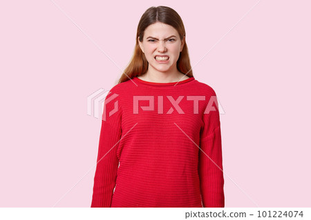 Dissatisfied dark haired woman clenches teeth, frowns face with annoyance, dressed in red sweater, models over rosy background. Grumpy irritated female model sees something negative and bad. Dissatisfied dark haired woman clenches teeth, frowns face with annoyance, dressed in red sweater, models over rosy background. Grumpy irritated female model sees something negative and bad. 101224074