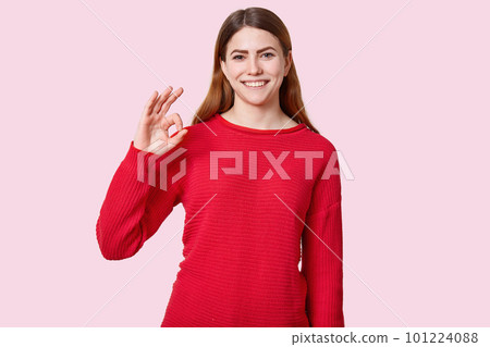 Happy pleased young female model smiles positively, shows okay sign, demonstrates approval, dressed in red clothes, isolated over rosy background. People, agreement and satisfaction concept. Happy pleased young female model smiles positively, shows okay sign, demonstrates approval, dressed in red clothes, isolated over rosy background. People, agreement and satisfaction concept. 101224088