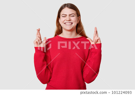 Indoor shot of pleasant looking dressed in casual red sweater, crosses fingers and has great hope for better, waits results of examination, posing in studio with cluosed eyes and toothy smile. 101224093