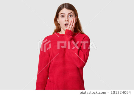 Emotive portrait of beautiful fashionable girl dressed red sweater posing over white background. Attractive female has astonished facial expression, keeps palm on cheek, stands with opened mouth. Emotive portrait of beautiful fashionable girl dressed red sweater posing over white background. Attractive female has astonished facial expression, keeps palm on cheek, stands with opened mouth. 101224094