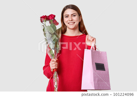 Joyful pretty woman with straight dark hair, smiles gently, holds roses and gift bag, dressed in red sweater, isolated over white background, recieves present on birthday. People and holiday concept Joyful pretty woman with straight dark hair, smiles gently, holds roses and gift bag, dressed in red sweater, isolated over white background, recieves present on birthday. People and holiday concept 101224096