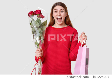 Shot of pleased overjoyed woman with dark hair, dressed in oversized red sweater, carries roses and gift bag, recieves package, isolated over white background. People, happiness, spring time Shot of pleased overjoyed woman with dark hair, dressed in oversized red sweater, carries roses and gift bag, recieves package, isolated over white background. People, happiness, spring time 101224097