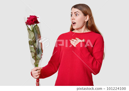 Scared surprised young European woman keeps hand on chest, looks with wonder at bouquet of red beautiful roses, wears casual jumper, revieves flowers from stranger, isolated over white background 101224100