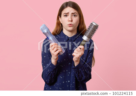 People, beauty, grooming concept. Displeased young woman pouts lips dressed in stylish shirt, poses over rosy background, holds hairspray and comb, comes to hairdresser for making hairstyle. 101224115