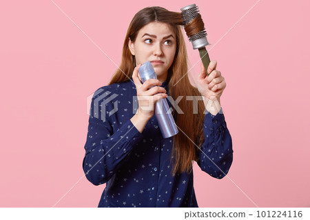 Teenage girl hurries for date, tries to make hairstyle, combs hair with hairbrush, uses hairspray, dressed in shirt, poses over rosy studio background. Pretty woman prepares for important meeting 101224116