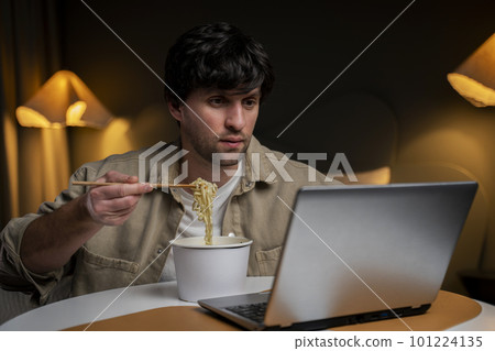 Business man eats Asian takeaway noodles while working at a computer laptop while sitting in the bedroom Business man eats Asian takeaway noodles while working at a computer laptop while sitting in the bedroom 101224135