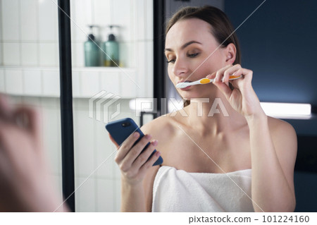 Young woman in bathroom brushing her teeth while reading messages on mobile phone 101224160