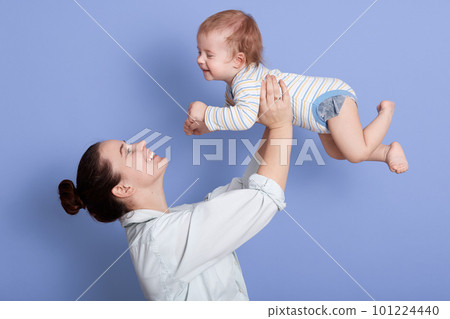 Portrait of happy mother and child together having fun isolated over blue background, attractive female with bun, dresses white shirt, infant wearing striped bodysuit. Mom and son, family concept. 101224440