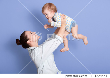 Side view of mid adult Caucasian mom lifting happy toddler son into air above head, attarctive mommy wearing white shirt, having dark hair with bun, happy child spending time with his beloved mom. 101224441