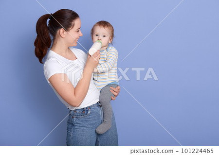 Horizontal indoor photo of cheerful attractive young brunette with ponytail, holding her child, feeding him, having pleasant facial expression, wearing casual clothes. Copyspace for advertisement. 101224465