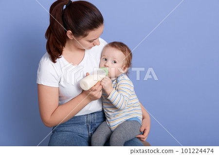 Image of caring attractive young mother sitting at chair, feeding her son with milk from bottle, wearing white t shirt and jeans, having ponytail, spending time with her child. Motherhood concept. Image of caring attractive young mother sitting at chair, feeding her son with milk from bottle, wearing white t shirt and jeans, having ponytail, spending time with her child. Motherhood concept. 101224474