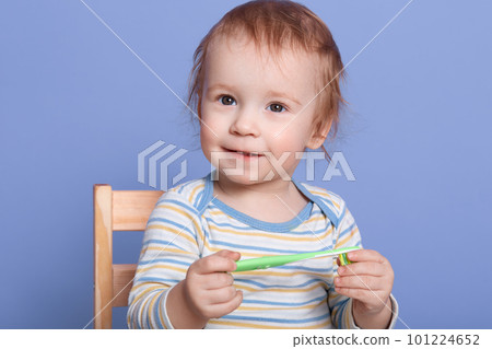 Little boy with toothbrush in hads, posing isolated over blue background, looking smiling at camera, child being interested, baby sitting on chair, wearing striped shirt, infant playing by himself. Little boy with toothbrush in hads, posing isolated over blue background, looking smiling at camera, child being interested, baby sitting on chair, wearing striped shirt, infant playing by himself. 101224652
