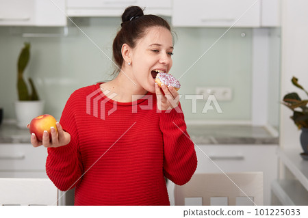 Pregnant woman in kitchen holding apple in hand and eating cake. Young attractive female decides to eat unhealthy food, looks happy, dressed casually, has hairstyle bun, posing in white kitchen. 101225033
