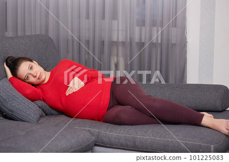 Dark haired attractive future mother lies on comfortable grey sofa, wearing warm bright red sweater and maroon leggings, holds her one hand on tummy, looks directly at camera, being peaceful. Dark haired attractive future mother lies on comfortable grey sofa, wearing warm bright red sweater and maroon leggings, holds her one hand on tummy, looks directly at camera, being peaceful. 101225083