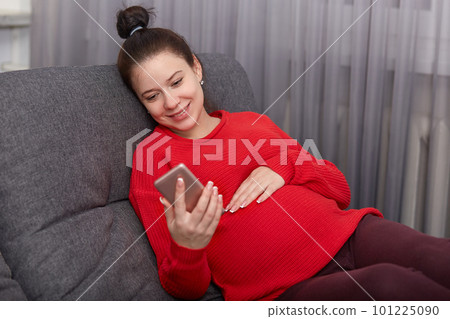 Pregnancy, leisure, technology concept. Happy smiling pregnant girl holds smart phone, wears red sweater, rests at couch, watches video for future mothers, makes shopping online, browses internet 101225090