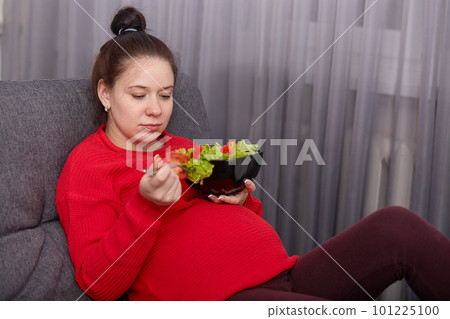 Image of adorable brunette pregnant lady enjoys eating fresh salad, has good appetite, being in modern apartment on sofa, dressed red sweater and leggins, taking care of her health during pregnancy. Image of adorable brunette pregnant lady enjoys eating fresh salad, has good appetite, being in modern apartment on sofa, dressed red sweater and leggins, taking care of her health during pregnancy. 101225100