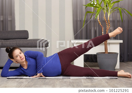 Healthy lifestyle during pregnancy. Pregnant woman lies on her hip and lifting her leg, has dark hair combed in knot, models posing while lying on floor and exercising, takes care of her health. Healthy lifestyle during pregnancy. Pregnant woman lies on her hip and lifting her leg, has dark hair combed in knot, models posing while lying on floor and exercising, takes care of her health. 101225291