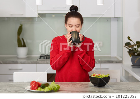 Close up portrait of good looking woman keeps cup of tea or coffee with both hands, drinks hot beverage, pregnant female poses against kitchen interior, enjoys calm atmosphere. Motherhood concept. Close up portrait of good looking woman keeps cup of tea or coffee with both hands, drinks hot beverage, pregnant female poses against kitchen interior, enjoys calm atmosphere. Motherhood concept. 101225352